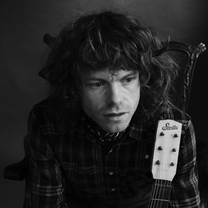 A black and white photo of Jesse Welles sitting in a vintage chair with his guitar. Welles stares off in a some what dissociative manner with his hair in disarray.