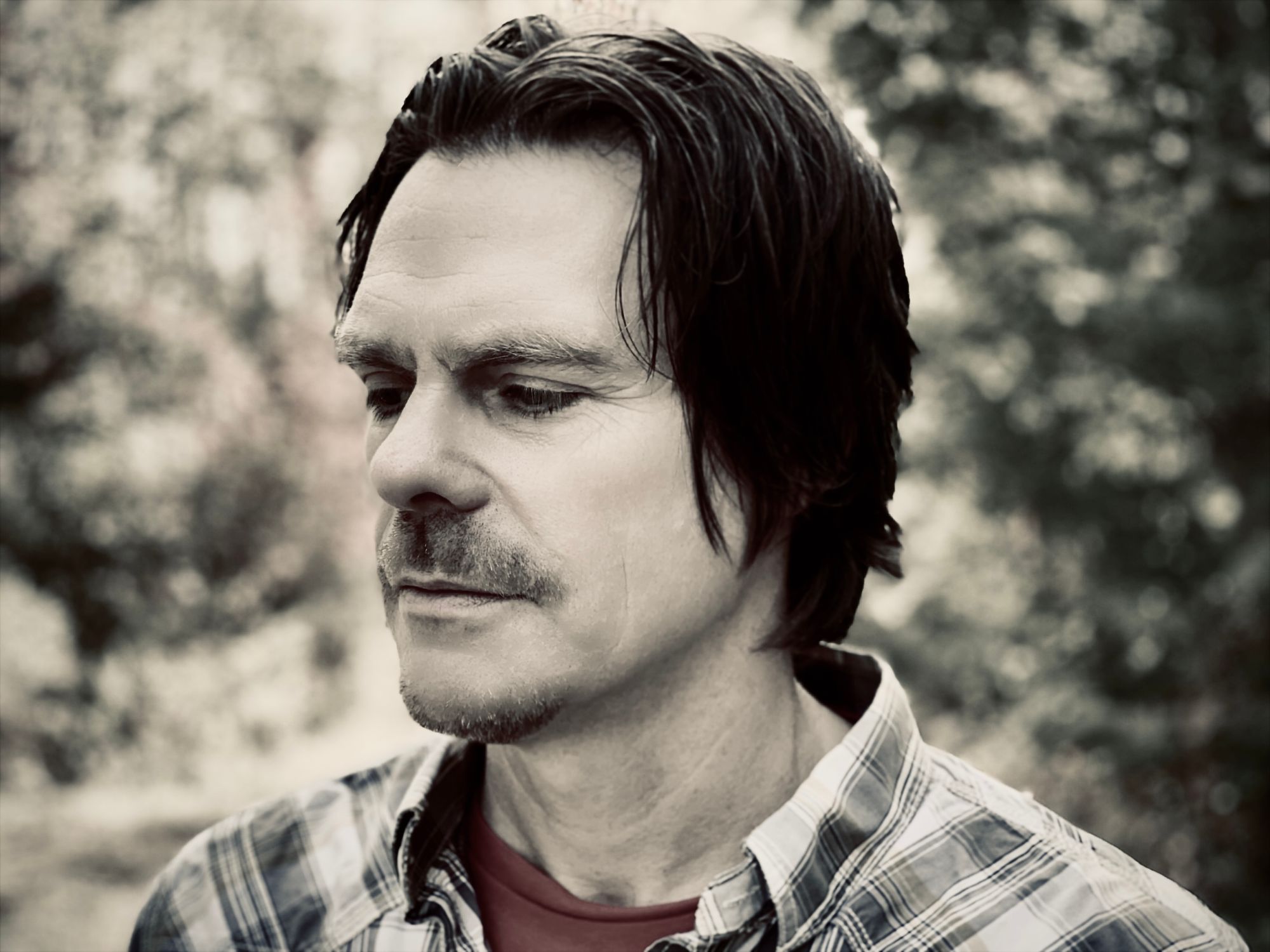 Desaturated, nearly black and white photo of Rick Sparkes with the exception of his red but muted t-shirt worn under his dress shirt. Sparkes stares at the ground in a contemplating manner with out of focus foliage in the background.